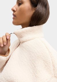 Kremowa polarowa kurtka z wysokim kołnierzem i zamkiem, prezentująca miękką teksturę i luźny krój. Akcentowana beżowym podszyciem.