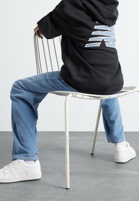 Persona in pantaloni di velluto a coste blu, sneakers bianche e felpa nera, seduta al contrario su una sedia di metallo bianca su un pavimento grigio.