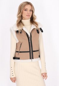 Tan vest met zwarte accenten, schapenvacht kraag, ritssluiting aan de voorkant, minimalistisch ontwerp en twee zijzakken. Model draagt een crèmekleurige ribbeltrui.