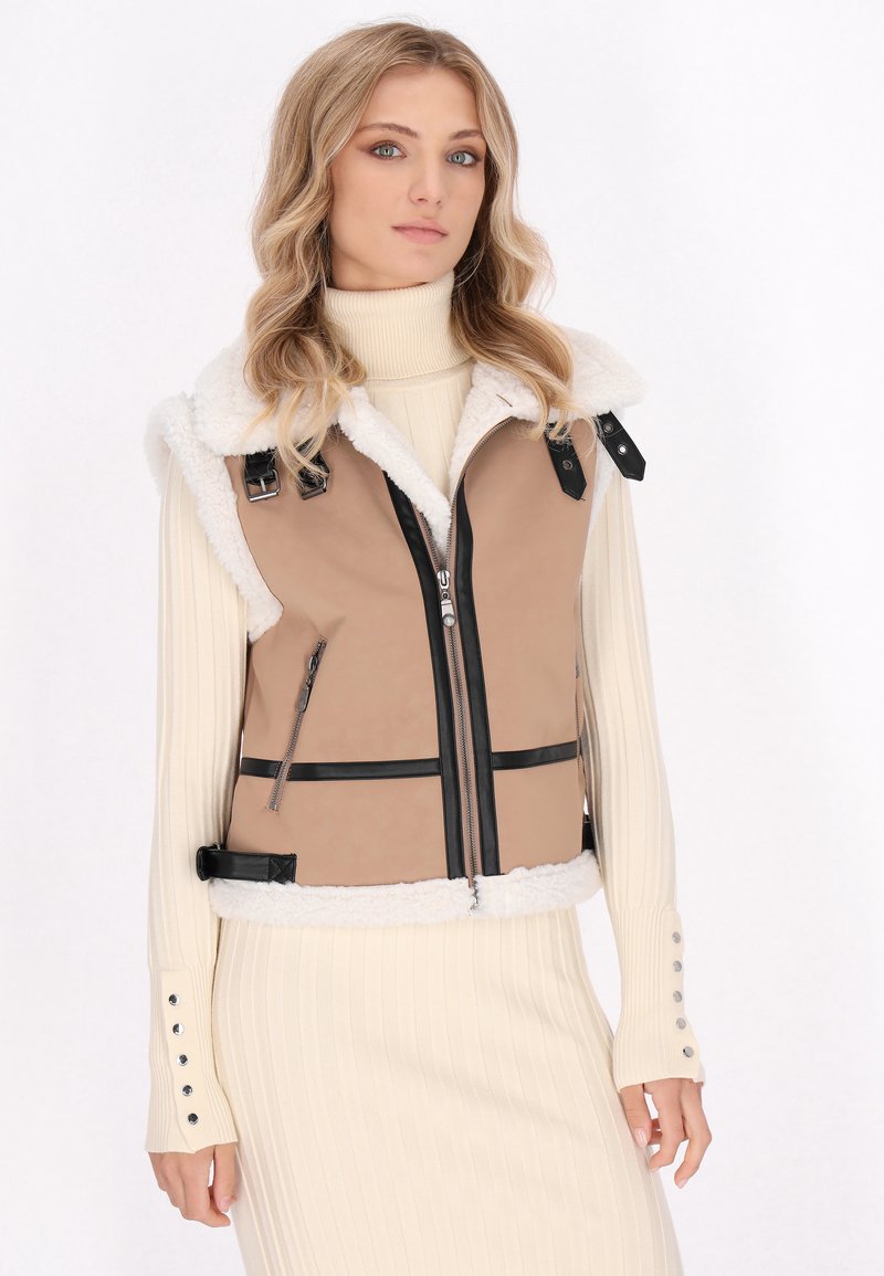 Tan vest met zwarte accenten, schapenvacht kraag, ritssluiting aan de voorkant, minimalistisch ontwerp en twee zijzakken. Model draagt een crèmekleurige ribbeltrui.
