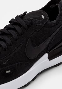 Sapatilha Nike preta com parte superior em malha e camurça, logotipo Swoosh preto, atacadores pretos e sola branca com detalhe de solado preto.