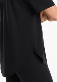 Black top made of smooth fabric, featuring a curved hem, short sleeves, and side slits for a relaxed fit.