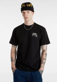 Camiseta de algodón negra con un pequeño logo blanco de Vans y un diseño gráfico. Combinada con jeans de mezclilla y un gorro de punto negro con llamas amarillas.