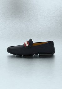 Mocassin en cuir noir avec détail de rayures rouges et blanches sur le dessus, bout arrondi et semelle en caoutchouc, présenté sur une surface gris clair.