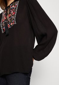 Svart blus med långa puffärmar och en färgglad broderad halsringning med fransdetaljer. Mjuk, texturerad tyg.