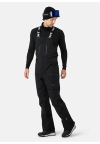 Zwarte sneeuw overalls met verstelbare bandjes, ritszakken en een taps toelopende pasvorm. Draagt een aansluitend zwart long-sleeve shirt en zwarte laarzen.