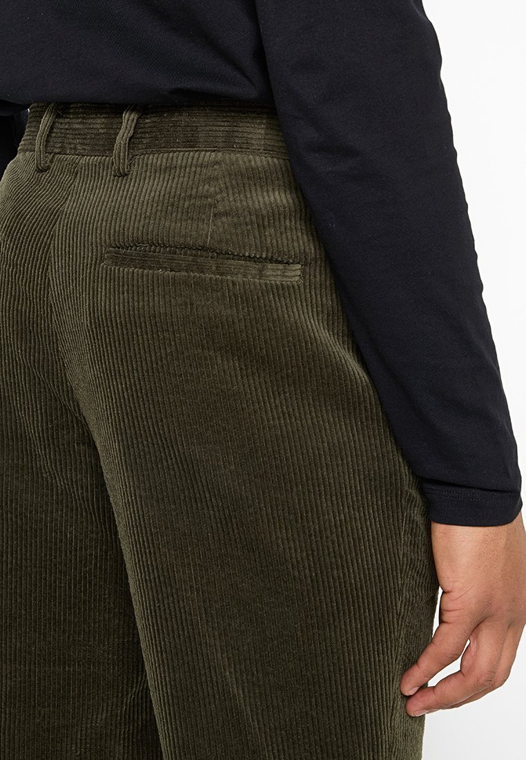 Olivgrüne Cordhosen mit strukturiertem Rillenmuster und einer Gesäßtasche, getragen mit einem schwarzen Langarmshirt.