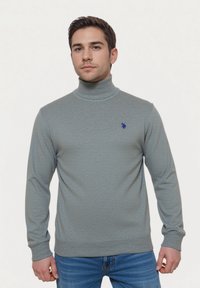 Hellgrauer Rollkragenpullover mit strukturiertem Muster, gerippten Bündchen und Saum. Verfügt über ein kleines blaues Logo auf der Brust. Getragen mit blauen Jeans.