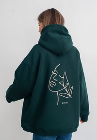 Groene hoodie van zacht materiaal met een grote grafische weergave van een gezicht en een plant lijntekening op de achterkant, met roze borduurdetails.