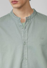 Lichtgroene button-up shirt met een bandkraag, voorzien van drie knopen en subtiele stikseldetails. Logo op de borst in lichtgrijs.