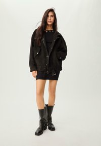 Jeune femme aux longs cheveux noirs portant une mini robe noire, une veste noire oversize avec des poches et des bottes en cuir noires mi-mollet, se tenant sur un fond blanc.