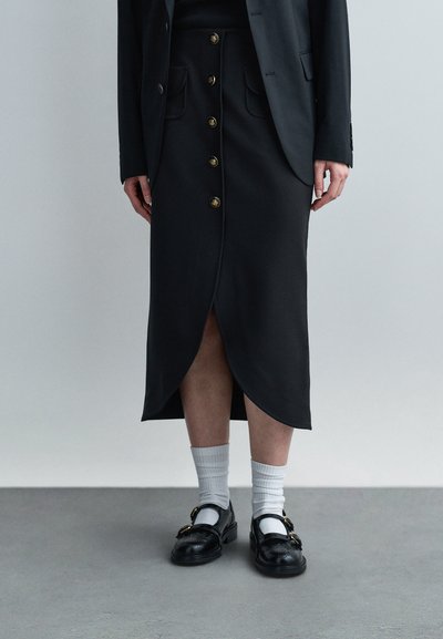 Person wearing black buttoned skirt with front slit, black blazer, white crew socks, and black buckle loafers standing on gray floor.