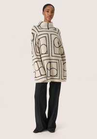 Beige oversized tröja med svarta geometriska mönster, hög krage och textilstruktur, i kombination med svarta vida byxor och mörka skor.