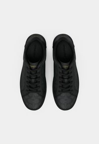 Schwarze Sneakers mit einer strukturierten Oberfläche und einem dezenten Muster. Schnürsenkel verlaufen durch Ösen, und ein goldenes Logo ist auf der Zunge zu sehen.