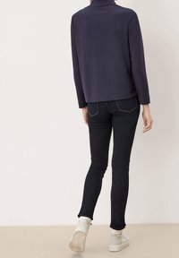 Pullover blu navy con colletto alto, abbinato a pantaloni in denim scuri. Il pullover è liscio, con una vestibilità rilassata e cuciture minime. Le calzature sono sneakers di colore chiaro.