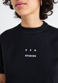 Zwarte katoenen t-shirt met een ronde halslijn, voorzien van witte bedrukte letters "C P H" boven "STUDIOS" in het midden. Glad, niet-geweven stof.