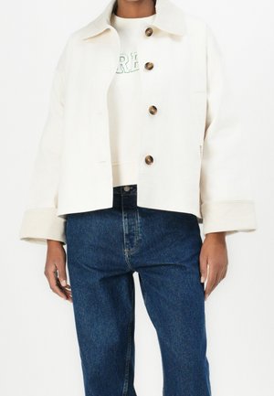 Person, die eine weiße Jacke mit Knöpfen und hochgekrempelten Ärmeln, ein weißes Hemd mit grünen Buchstaben und blaue Jeans vor schlichtem Hintergrund trägt.