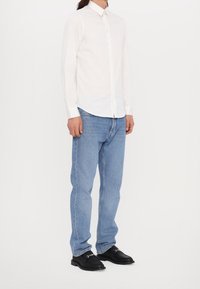 Homme portant une chemise blanche à manches longues, un jean bleu clair et des chaussures noires, debout devant un fond blanc uni.