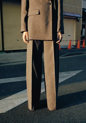 Oseba stoji na cesti v rjavem po meri krojenem kostimu z širokimi hlačnicami, blizu oranžnih prometnih stožcev in zaprtih vrat trgovine.