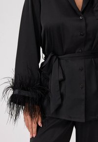 Czarna satynowa bluzka z długimi rękawami, z piórami wykończonymi przy mankietach oraz związaną w talii paskiem. Guziki zdobią przód.