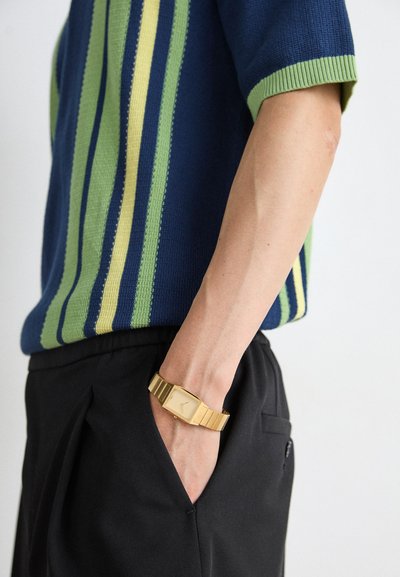 Knit short-sleeve sweater in blue with vertical green and yellow stripes; paired with black pants and a gold rectangular watch on the wrist.