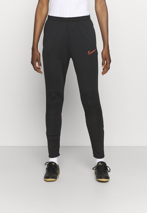 Osoba má na sobě černé sportovní kalhoty Nike s oranžovým logem swoosh, bílé ponožky a černé sportovní boty Adidas.