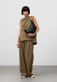 Borsa a mezzaluna in pelle nera con silhouette curvata, hardware dorato e manico corto, tenuta da una persona in un outfit verde oliva.