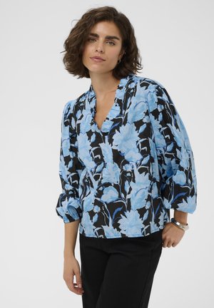 Žena s krátkymi kučeravými vlasmi oblečená v čiernej blúzke s jasným modrým kvetinovým vzorom a čiernych nohaviciach, pózujúca na jednoduchom pozadí.