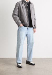 Grå corduroy bomberjakke med elastiske manchetter, sort polo, lyseblå lige ben jeans og sorte sko med hvide detaljer.
