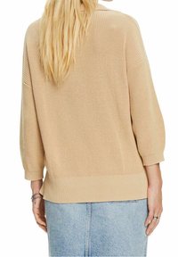 Femme aux cheveux longs et blonds portant un pull en maille beige à manches trois-quarts et une jupe en denim bleu clair, vue de dos.