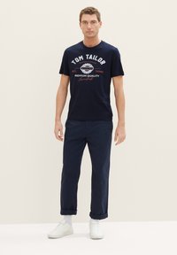 Marineblaue Baumwoll-T-Shirt mit weißem und rotem Logo-Druck, kombiniert mit marineblauen Hosen und weißen Sneakern. Einfaches, lässiges Design.