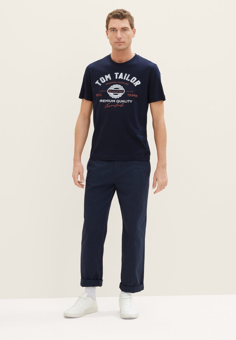 Marineblaue Baumwoll-T-Shirt mit weißem und rotem Logo-Druck, kombiniert mit marineblauen Hosen und weißen Sneakern. Einfaches, lässiges Design.