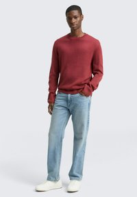Roter Strickpullover mit Rundhalsausschnitt, kombiniert mit hellblauen Jeans. Das Model steht mit entspannter Haltung und trägt weiße Turnschuhe.