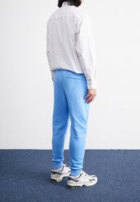 Person som står med ryggen mot kameran, klädd i en randig skjorta och blå mjukisbyxor, på en blå matta i ett enkelt vitt rum.