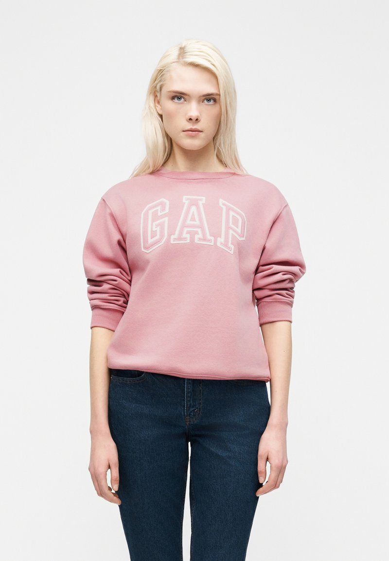 Ξανθιά γυναίκα που φοράει ροζ φούτερ GAP και σκούρο μπλε τζιν, στέκεται μπροστά από ένα μονόχρωμο λευκό φόντο με ουδέτερη έκφραση.