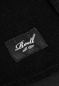 Schwarzes Gewebelabel mit dem Text "Reell" in weißer Schreibschrift und "est. 1997," auf einem strukturierten schwarzen Material Hintergrund gestickt.