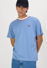 Camiseta de manga corta azul claro con cuello de canalé rojo y blanco y pequeño logotipo en el pecho. Ajuste informal, material de algodón.