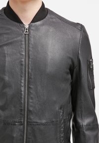 Giacca in pelle nera perforata con zip frontale, colletto a coste nero e tasca con zip sulla parte superiore del braccio sinistro.