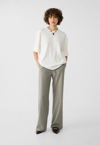 T-shirt bianco oversize, pantaloni grigi a vita alta e scarpe nere con punta. Presenta una collana con ciondolo a forma di cuore e una silhouette rilassata.