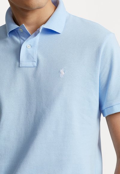 Gaišs zils polo krekls, izgatavots no teksturēta auduma, ar klasisku apkakli, trim pogām un baltu izšūtu logo kreisajā krūtī.