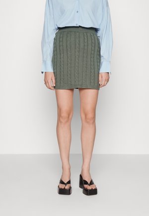 Femme portant une mini-jupe verte olive en tricot torsadé, une chemise bleu clair boutonnée et des sandales compensées noires, debout devant un fond uni.