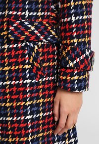 Vlněný kabát s houndstooth vzorem v červené, bílé, žluté a modré na tmavě námořnickém pozadí, s buttonem na manžetách a boční kapsou.