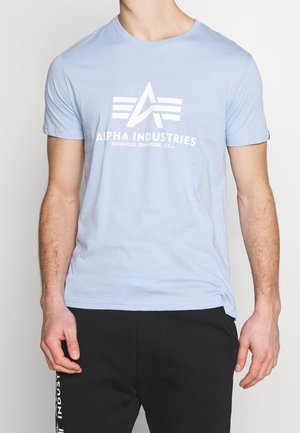Mężczyzna w jasnoniebieskiej koszulce Alpha Industries z logo i napisem, w połączeniu z czarnymi spodniami z białym napisem na udzie.