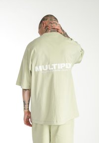Camiseta de corte holgado y manga corta en algodón verde claro. Presenta la palabra "MULTIPLY" impresa en texto blanco en negrita en la parte posterior. Textura suave.
