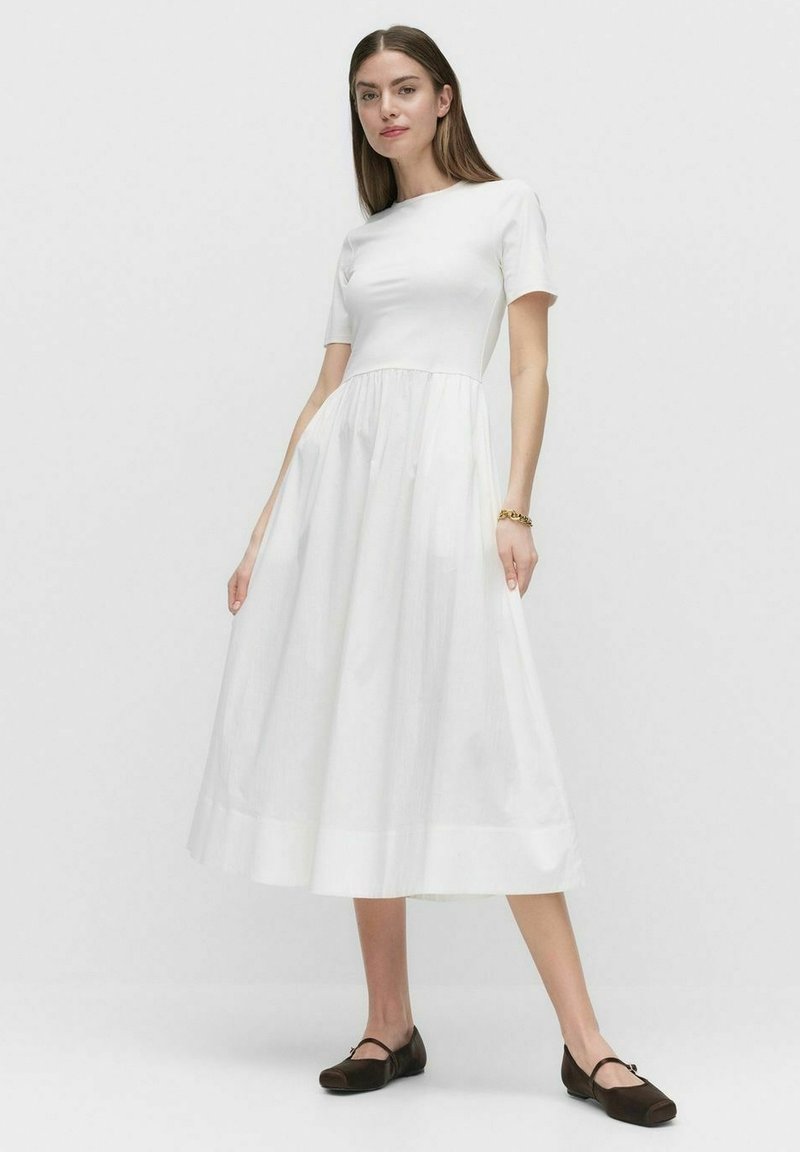 Vit midiklänning med korta ärmar, figursydd bodice och en utsvängd kjol. Snygg textur med en lätt glans, kombinerad med mörka balettklackar.
