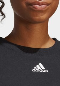 Svart sweatshirt med ribbat rundhals och vit Adidas-logotyp på bröstet. Slät tygstruktur, nära vy.