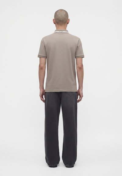 Homme à la tête rasée portant un polo beige à manches courtes et un jean ample gris foncé, debout de dos sur un fond uni.