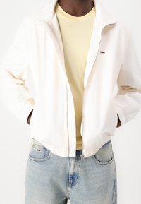 Lightweight white zip-up jacket with elastic cuffs, worn over a pale yellow T-shirt. Light blue jeans with a small logo on the pocket.