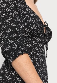 Vestido floral preto com pequenas flores brancas, mangas bufantes com punhos elásticos e um detalhe de laço no decote. Textura suave do tecido.