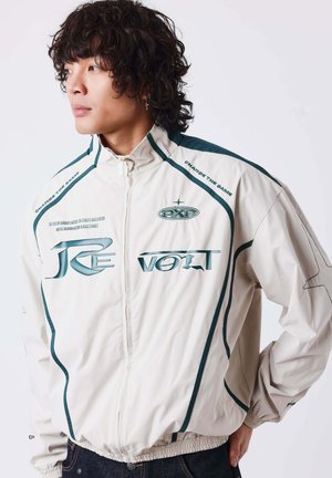 Homme aux cheveux bouclés portant une veste blanche zippée avec des accents vert foncé et du texte, debout, une main dans la poche, regardant vers la gauche.